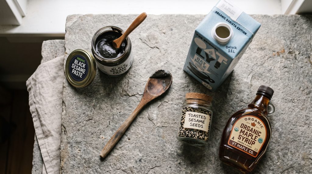 Black Sesame Latte: Easy 5-Minute Calming Evening Ritual 5 Top-down flat lay of black sesame paste, oat milk carton, maple syrup, and toasted sesame seeds on a stone countertop.