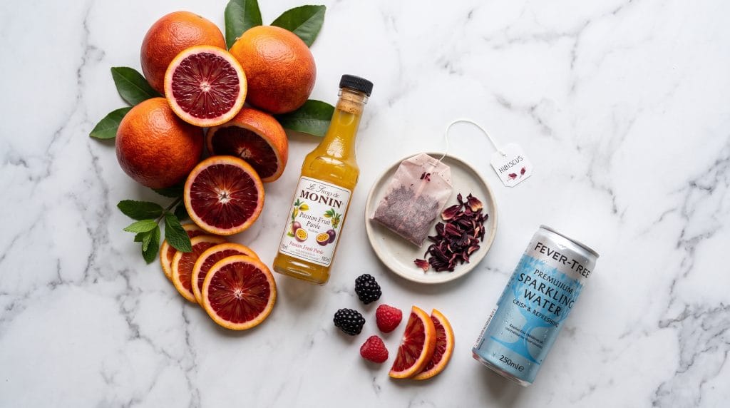 Sarti Spritz Mocktail: Easy 5-Minute Non-Alcoholic Drink 4 Flat lay of fresh blood oranges, passion fruit puree, hibiscus tea, and sparkling water on a marble surface.