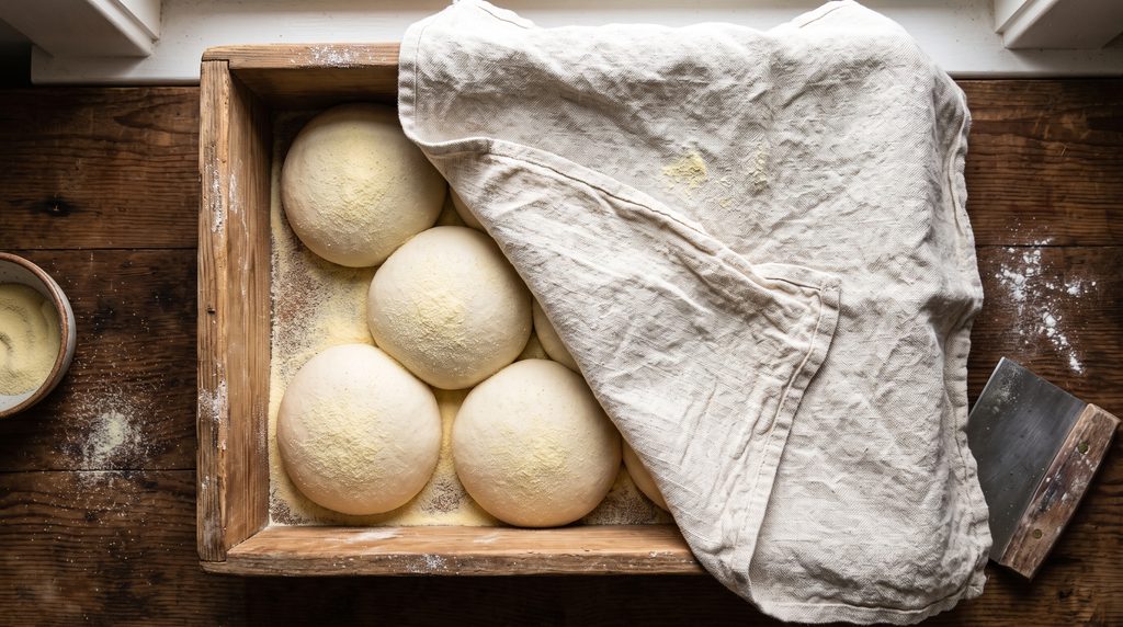 Overhead flat lay of pizza dough balls proofing on a floured wooden tray with linen cloth.